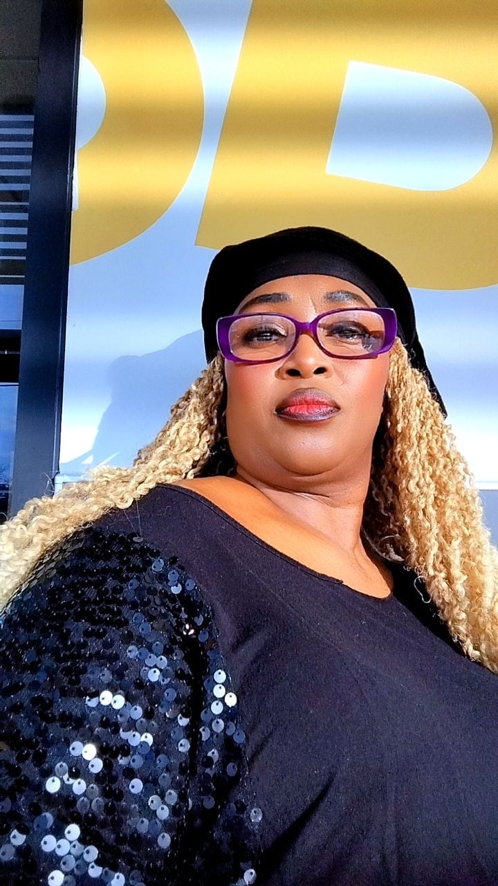 Person wearing purple glasses and black sequined jacket posing in front of gold signage outdoors