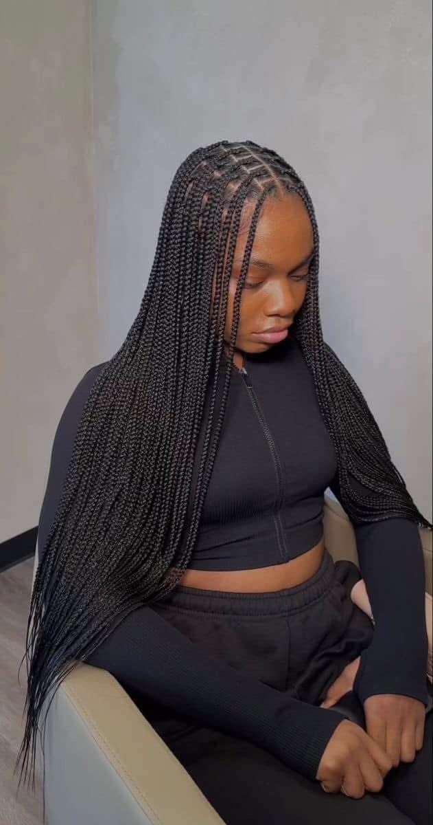 Woman with long box braids and cornrow pattern hairstyle, wearing black crop top and pants, seated indoors