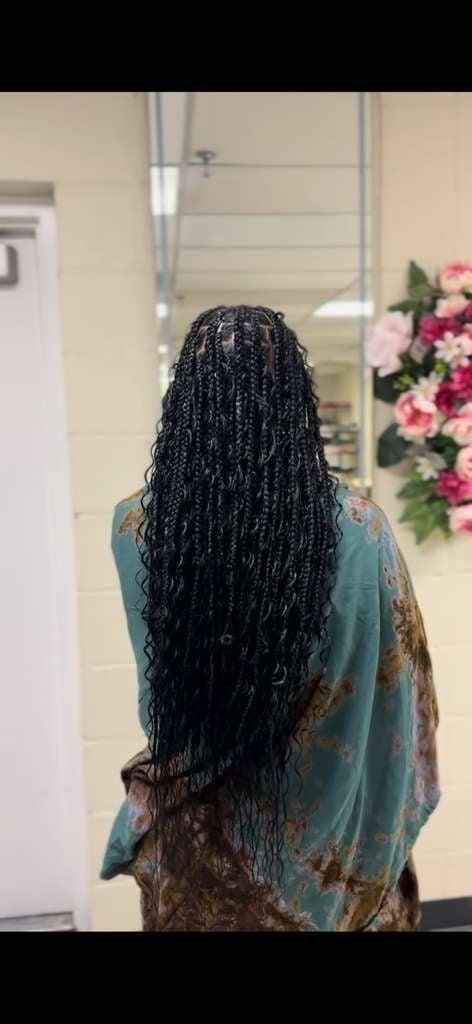 Person viewed from behind with long black braided hair, wearing a teal and gold patterned garment, standing in a bathroom with a mirror and pink flowers visible