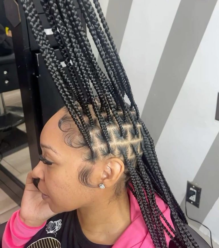 Profile view of person with long black box braids styled upward, showing scalp tattoo patterns and wearing pink and black clothing